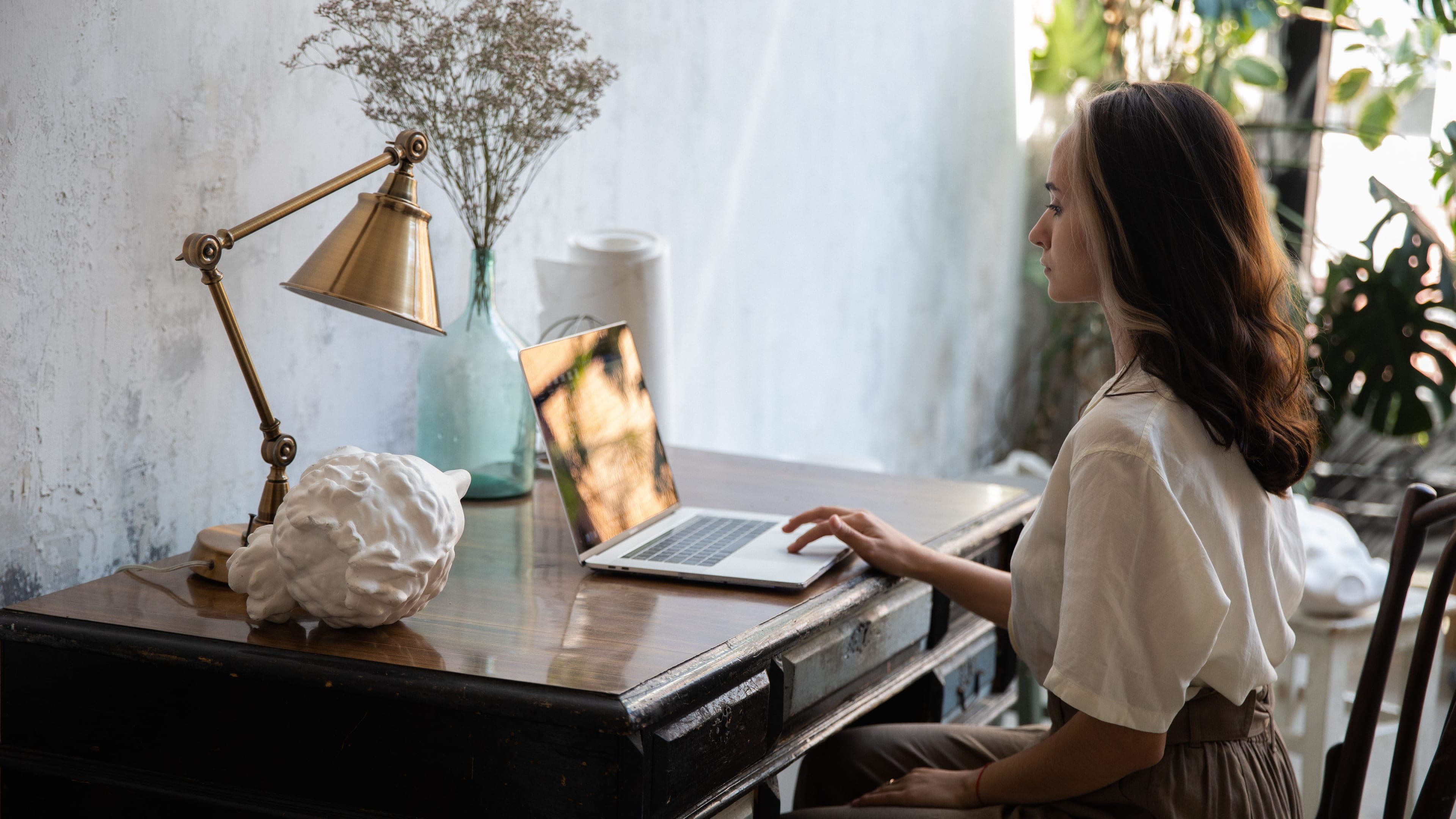 Eine Frau arbeitet an einem alten Holzschreibtisch mit Messinglampe und Trockenblumen am Laptop, hinter ihr stehen Pflanzen im Licht.