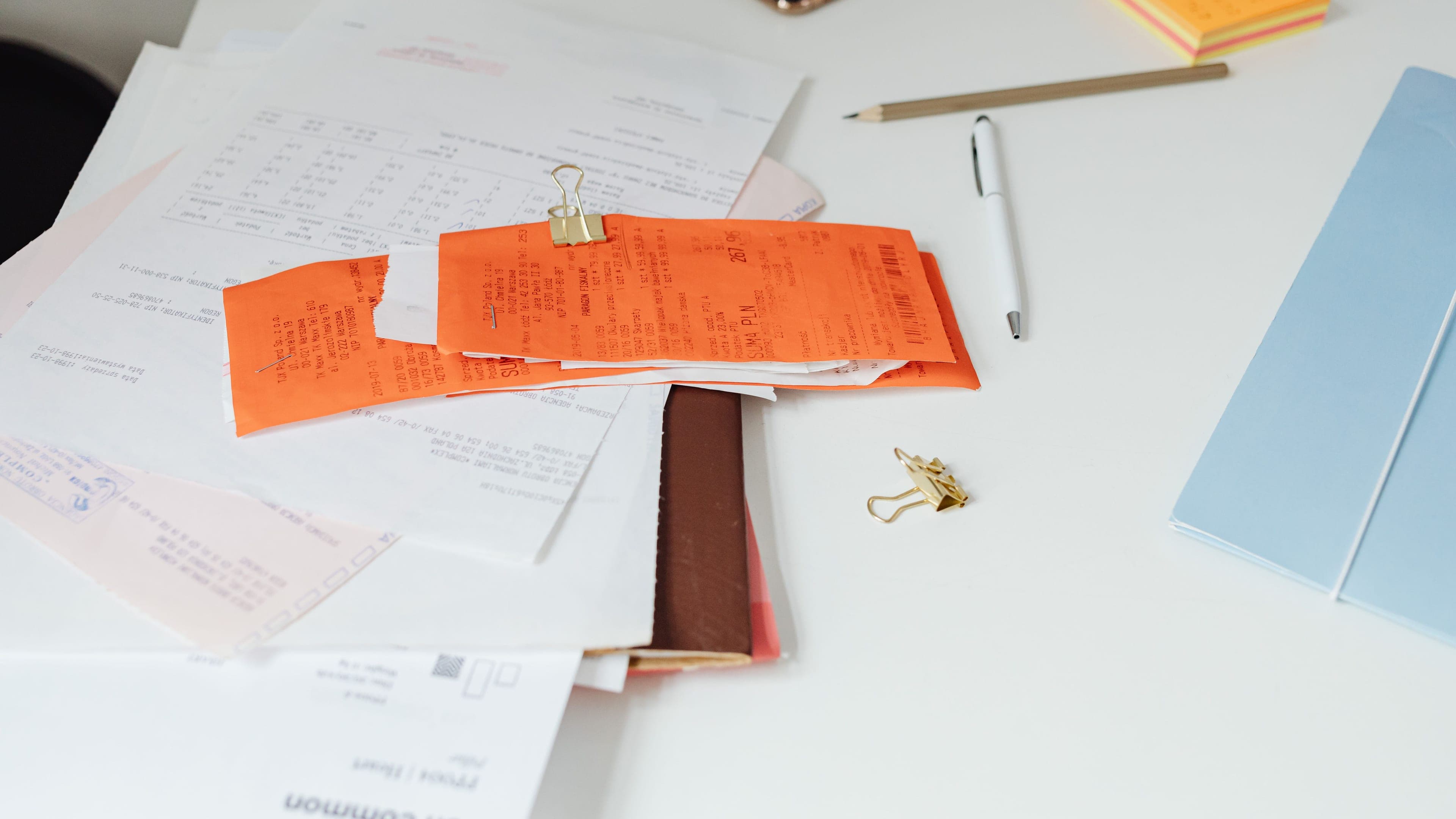 Papierrechnungen und orange Belege mit einer Klammer zusammengehalten auf einem weißen Schreibtisch, daneben ein Stift und eine blaue Mappe.