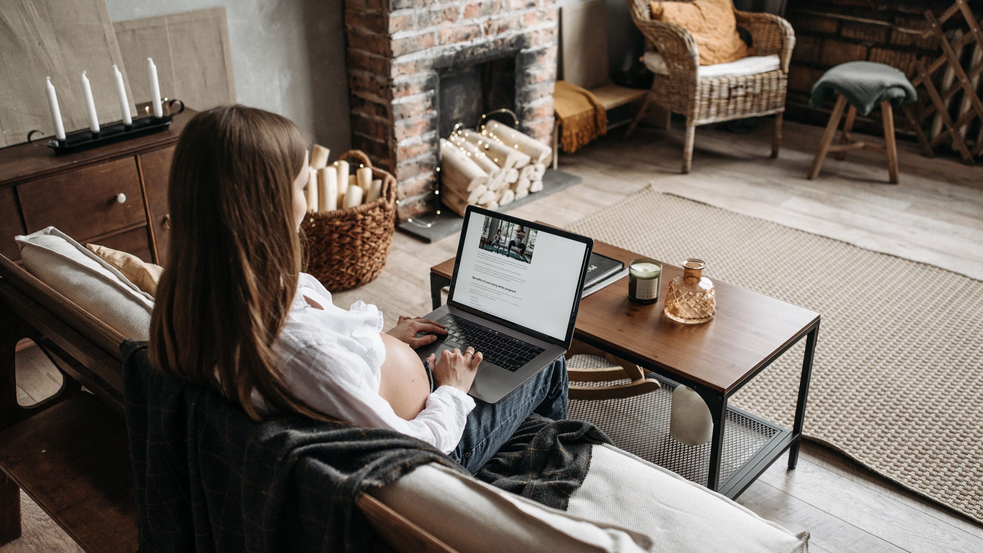 Eine schwangere Frau sitzt in einem warmen Wohnzimmer auf dem Sofa und arbeitet an einem Laptop auf einem kleinen Holztisch.